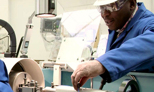 Three course students in blue coats stood around machine while an anonymous hand adjusts it