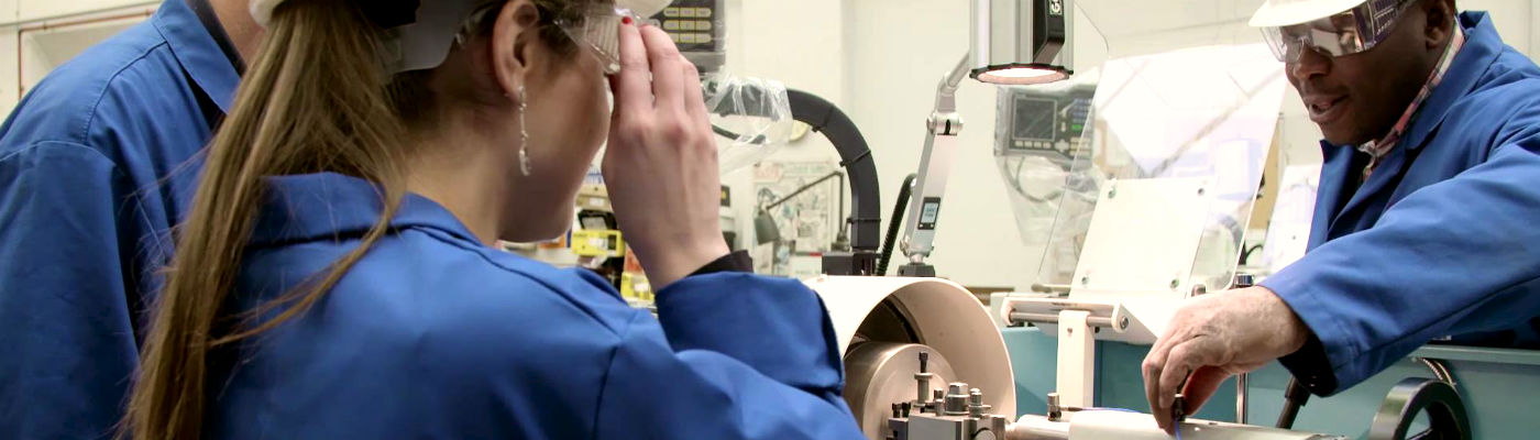 Three course students in blue coats stood around machine while an anonymous hand adjusts it